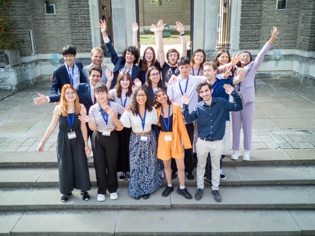 18 scholars smiling and waving their arms grouped together on some stone steps