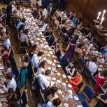 Overhead shot of people sat at 3 long tables in a row and another running perpendicular to these at the top of a large, old hall - Clare Great Hall. 