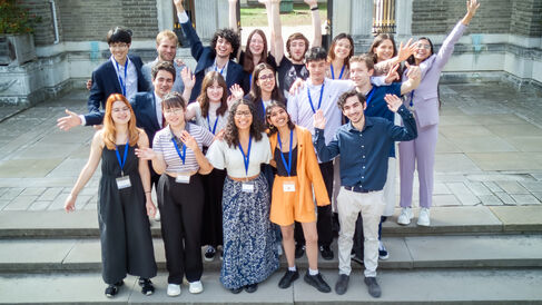 18 scholars smiling and waving their arms grouped together on some stone steps